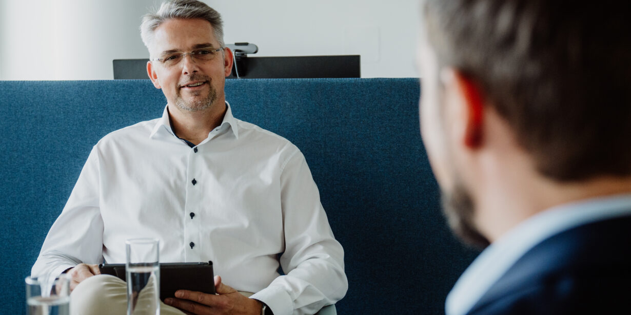 York Ambros frontal im weißen Hemd, wie er einer anderen Person freundlich etwas erklärt